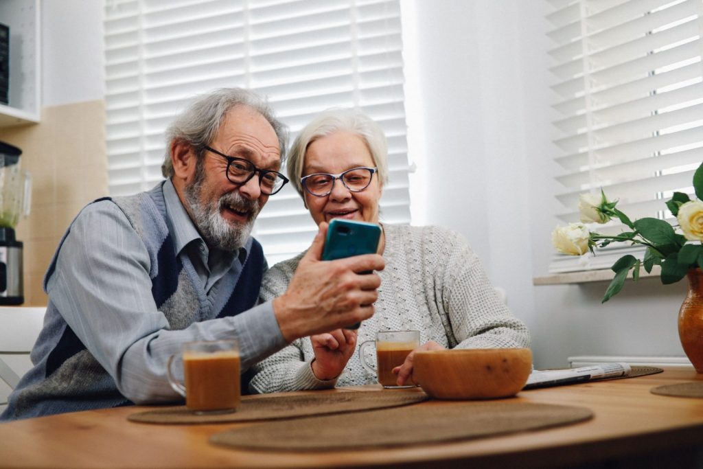 Happy senior couple sitting at a table, enjoying coffee and looking at a smartphone together in their cozy kitchen.